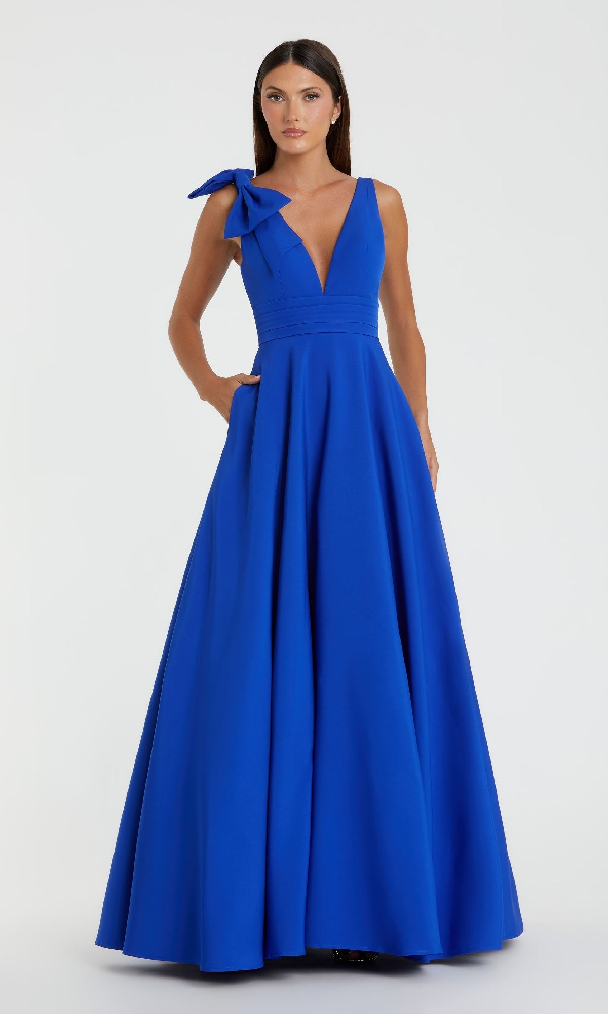 A woman in the Mac Duggal 49972 Shoulder-Bow Long Formal Dress, featuring a sleeveless royal blue design with a deep V-neck and large shoulder bow, stands against a plain light background.